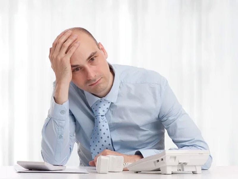 A man in a light blue shirt and tie sits at a desk with a hand on his head, appearing frustrated. A phone and papers are on the desk.