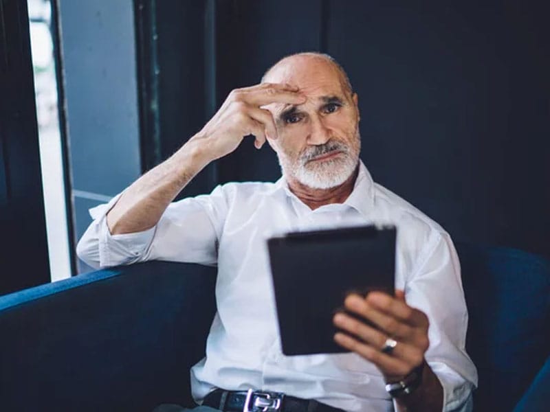 An elderly man with a beard sits on a blue chair, holding a tablet and touching his forehead with a thoughtful expression.